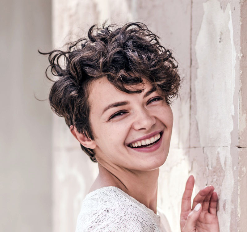 Woman smiling against a white wall with a tousled asymmetrical curly pixie cut