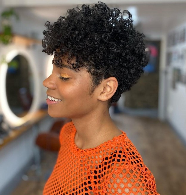 Side profile of a woman wearing an orange mesh top with a sculpted dark curly pixie